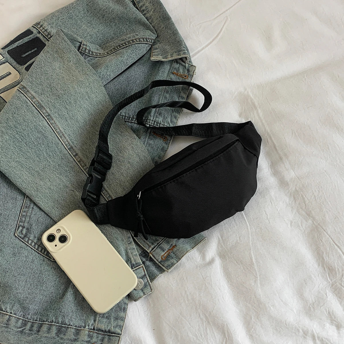A black nylon crossbody sling bag is placed on a light gray denim jacket and a white shirt.