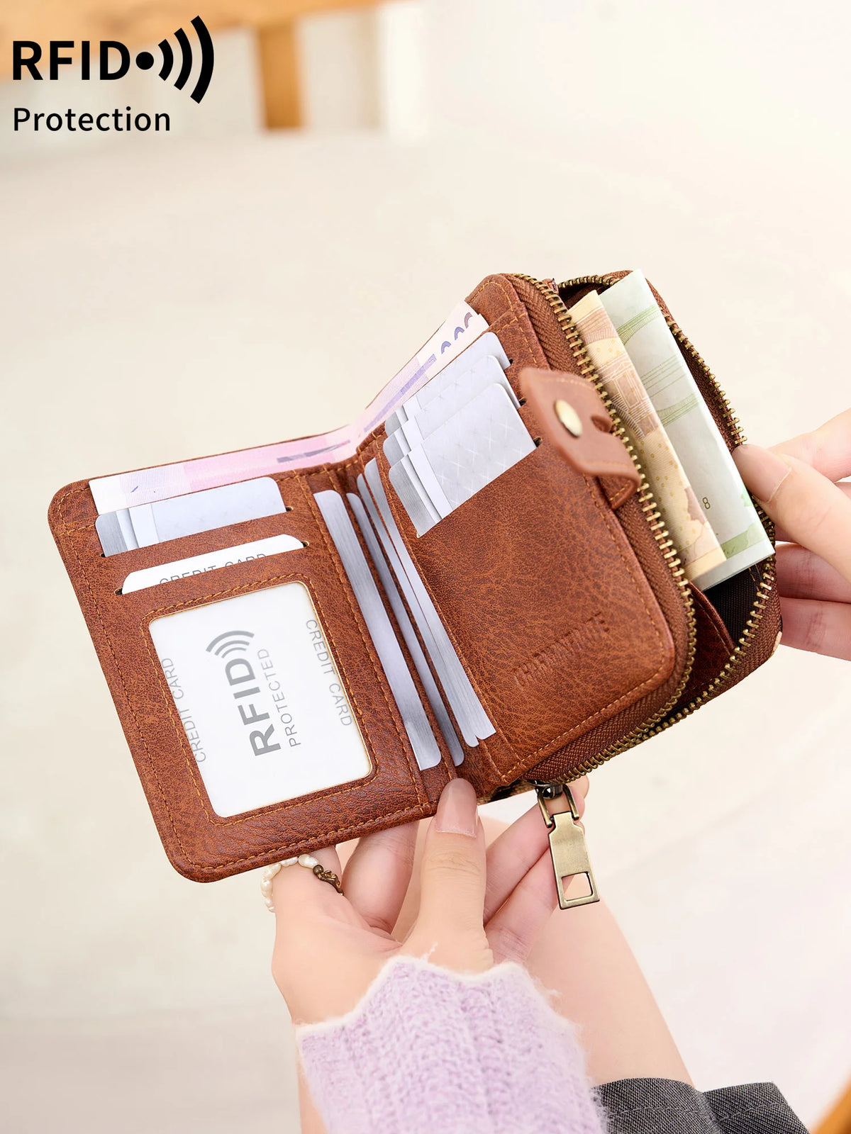 A brown RFID protected bi-fold wallet with a zipper closure is shown being held by a hand.