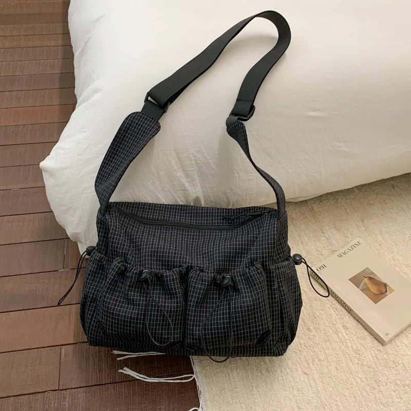 A black plaid nylon drawstring tote bag with a black strap resting on a white pillow next to a wooden surface.