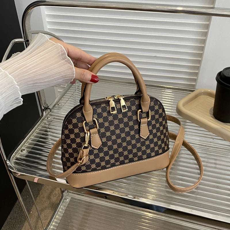 A tan fashion shell shoulder crossbody bag with gold hardware resting on a clear acrylic shelf.