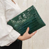 A hand holds a dark green alligator print clutch bag against a light background.