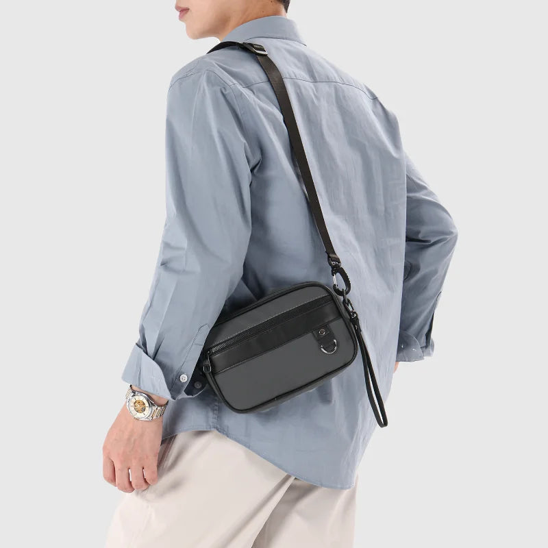 Una persona vistiendo una camisa azul claro y pantalones beige lleva un bolso cruz de cuero sintético negro.