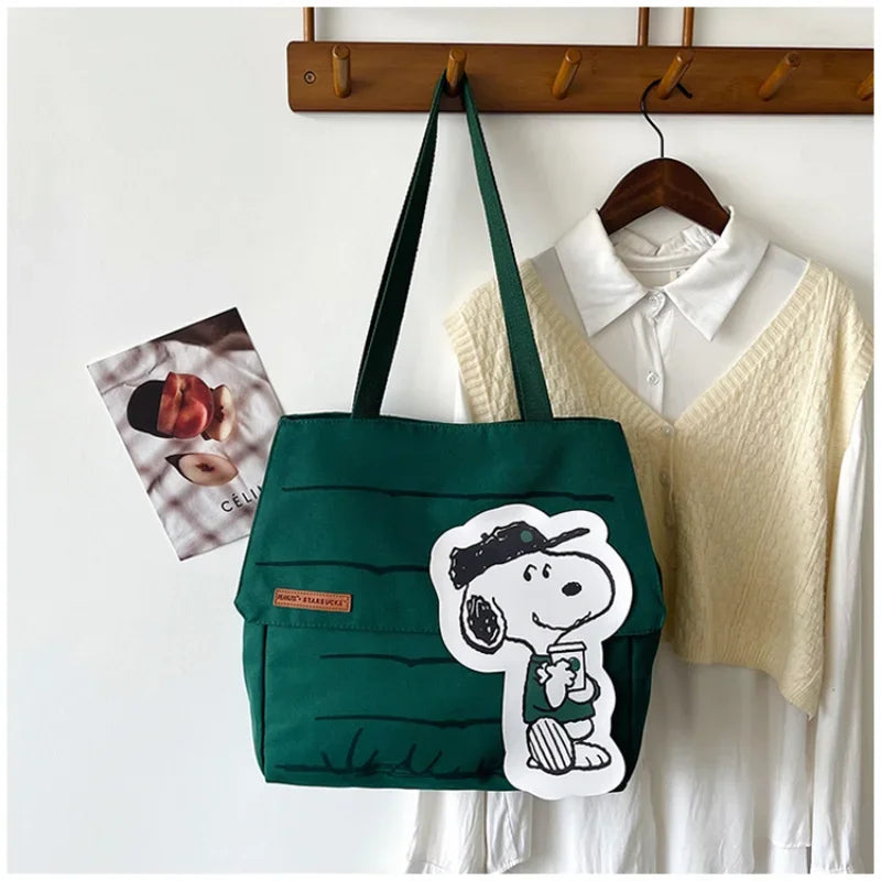 A dark green canvas tote bag featuring a white Snoopy design, hanging on a wooden rack.