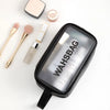 A clear waterproof travel toiletry bag with a black lid and a black strap, next to makeup brushes and powder.