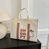 A beige canvas shoulder bag featuring a Snoopy cartoon design with brown stripes, resting on a dark surface.