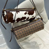 A brown and white cow print cylinder shoulder bag is displayed hanging on a leather strap against a light-colored sofa.