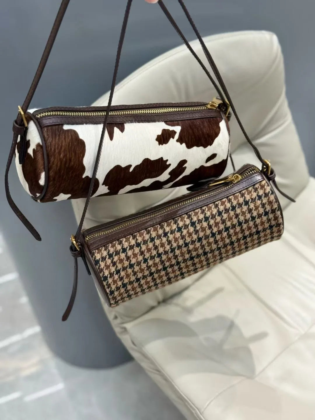 A brown and white cow print cylinder shoulder bag is displayed hanging on a leather strap against a light-colored sofa.