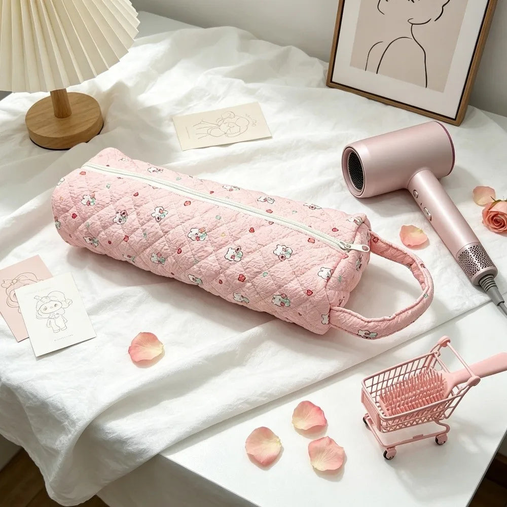 A pink quilted storage bag is displayed on a white surface next to a pink hair dryer and some rose petals.