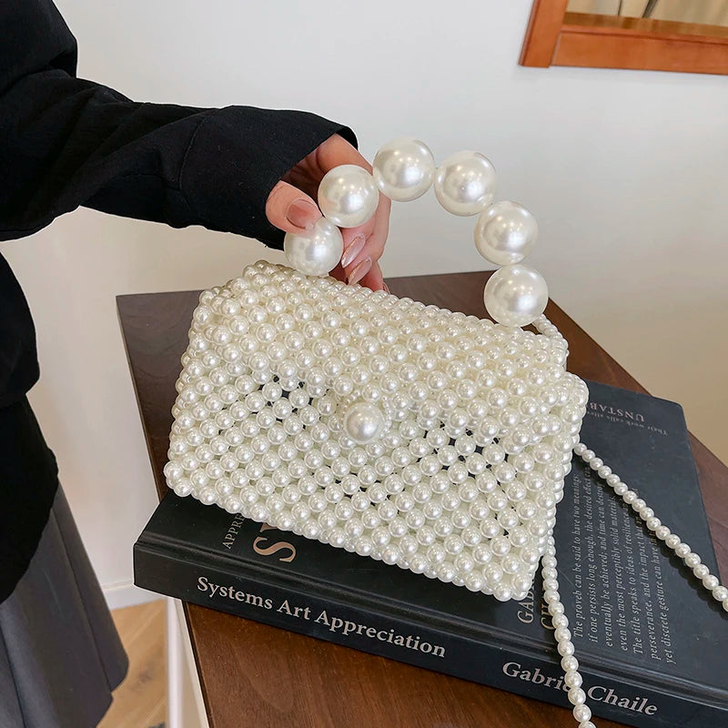 A hand holds a handmade pearl beaded clutch bag with a long pearl string strap on a dark book.