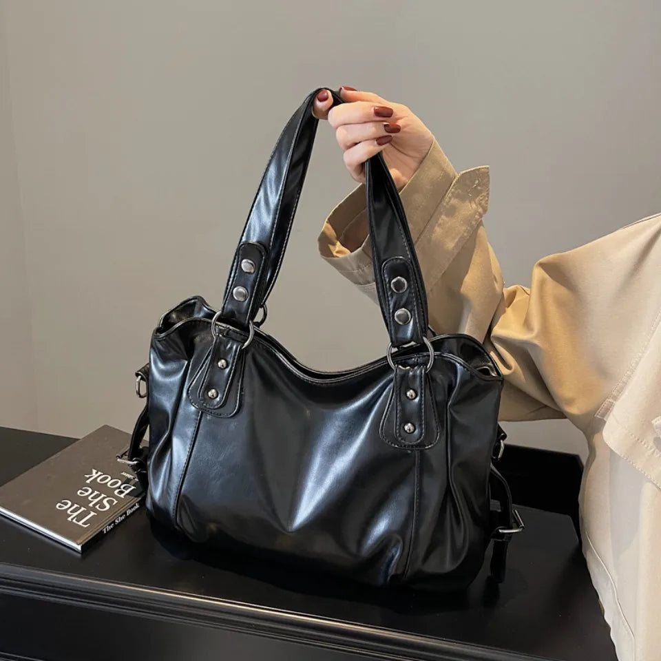 A hand holds a black leather tote bag with a strap against a light background.