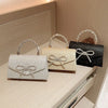 Tres bolsos de mano tipo lazo brillante para la noche en plata, beige y negro, exhibidos sobre una superficie clara.