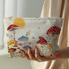 A beige makeup bag with a fairytale mushroom and butterfly print is held by a hand against a light background.