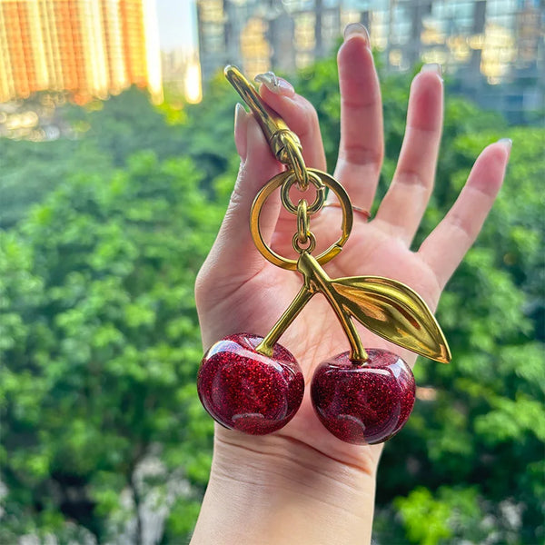 A hand holds a gold-toned crystal fruit charm keychain with a shiny red apple pendant against a blurred green outdoor backgro