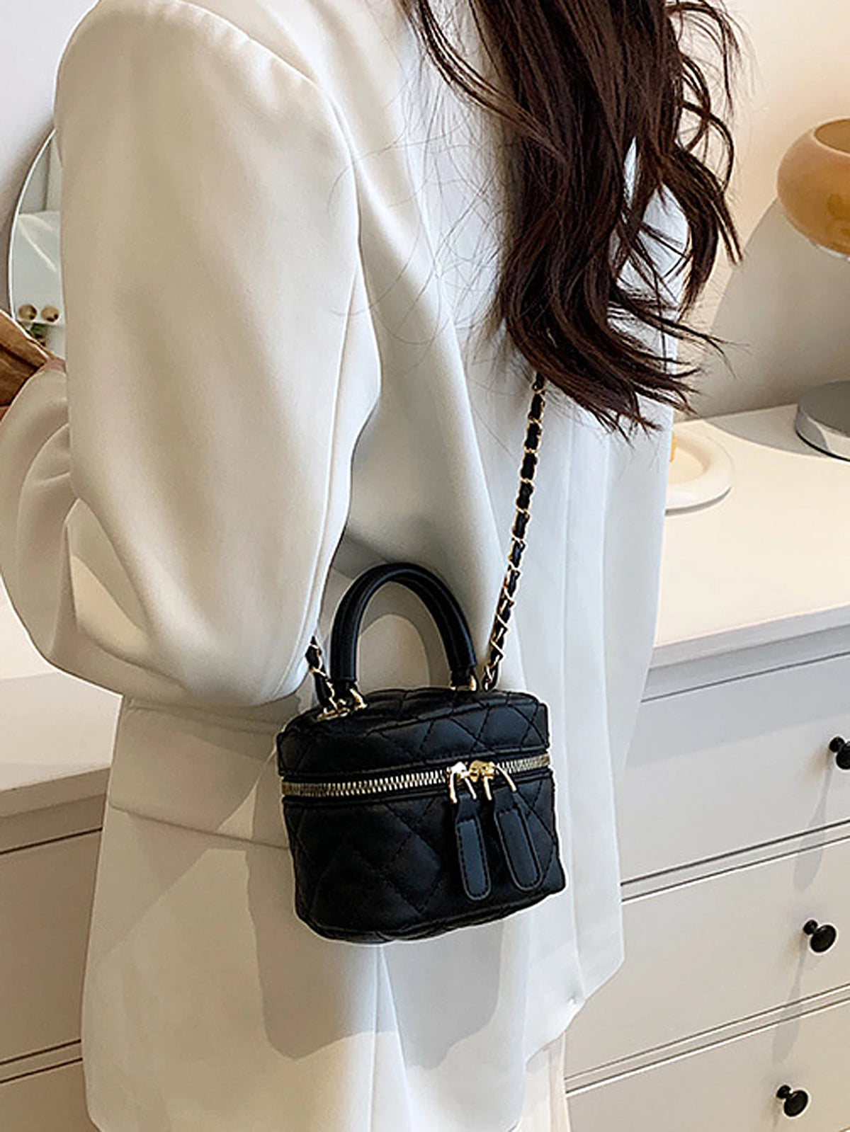 Gros plan d'une femme tenant un mini sac bandoulière noir matelassé avec une chaîne dorée, porté sous un blazer blanc.