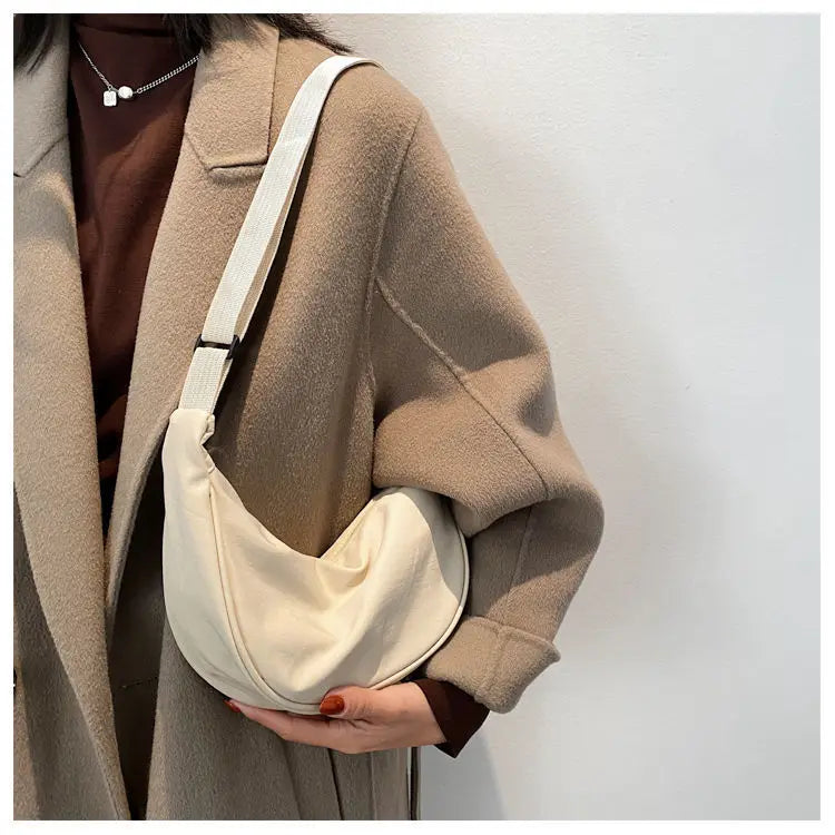 A woman wearing a beige coat is holding a cream-colored sling crossbody bag.
