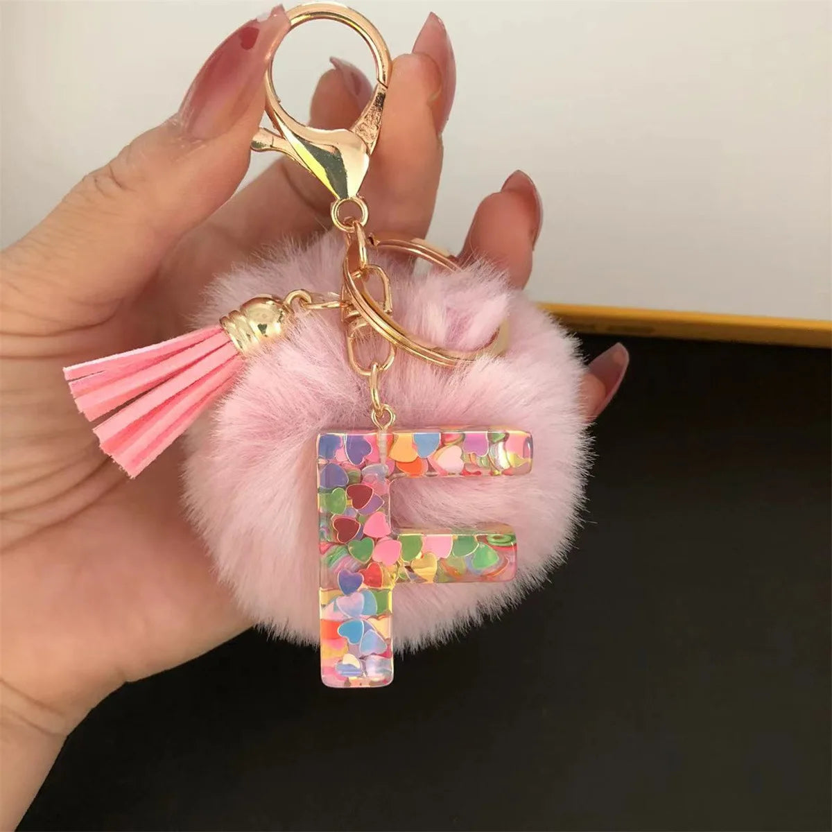 A hand holds a custom initial letter keychain featuring a shiny resin pendant with a pink alphabet charm and a fluffy pink po