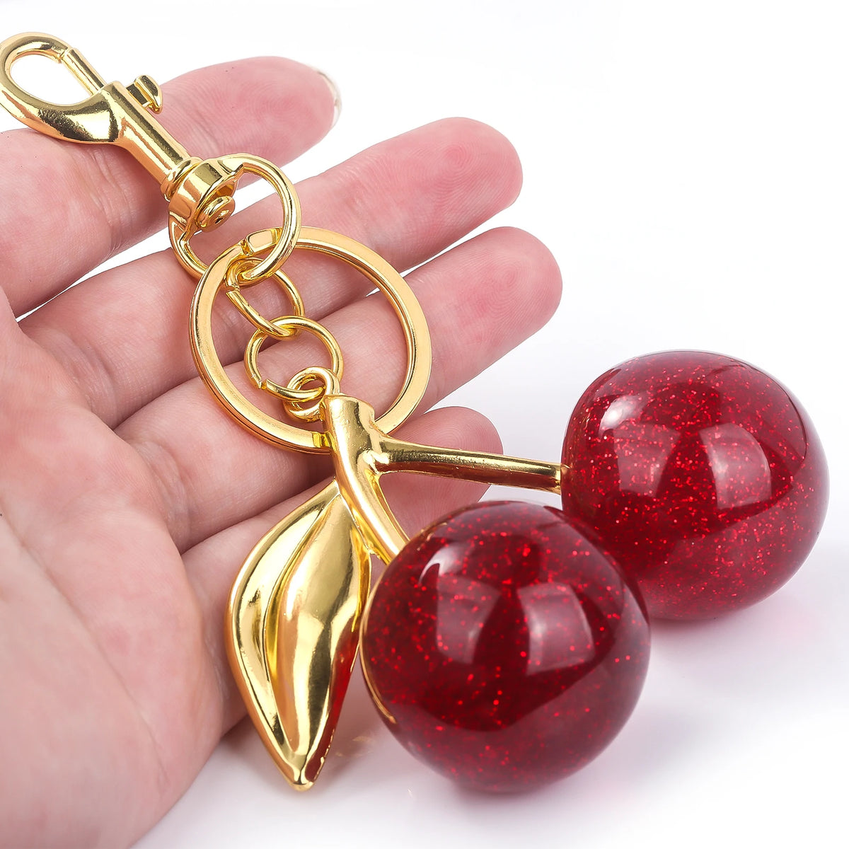 A gold-colored keychain featuring a red, glittery crystal cherry fruit charm held in a hand.