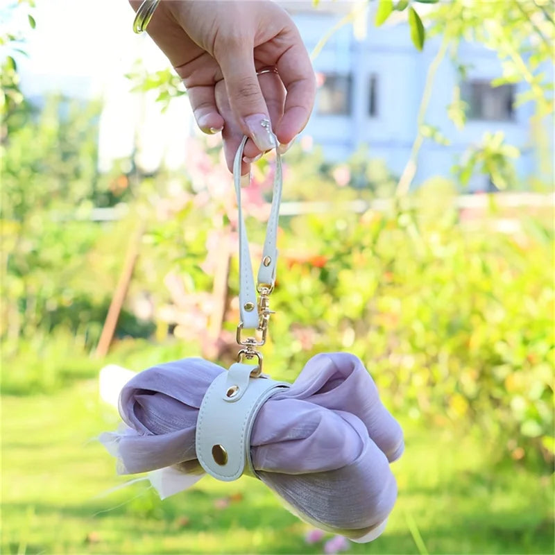 A hand holds a vintage leather glove holder strap with a white buckle against a blurred green outdoor background.