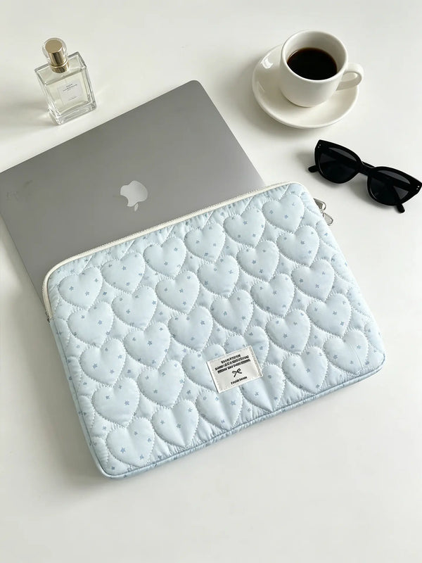 A light blue quilted laptop sleeve is placed next to a silver laptop and sunglasses on a white surface.