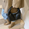 A close-up of a black woven embossed hobo shoulder bag with a top handle being held by a person wearing a beige jacket and je