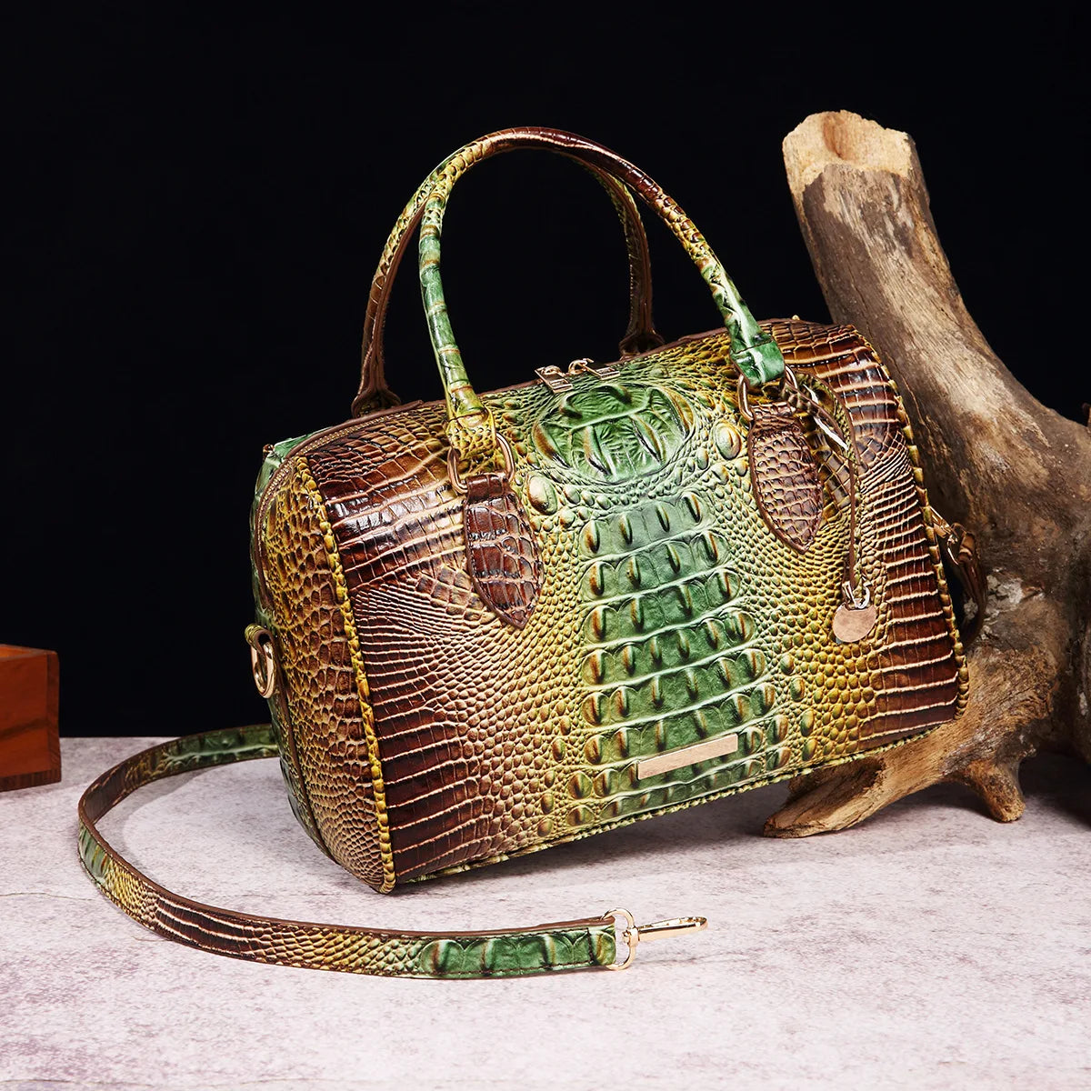 A luxury crocodile pattern crossbody tote bag with leather handles rests on a light surface next to a piece of driftwood.