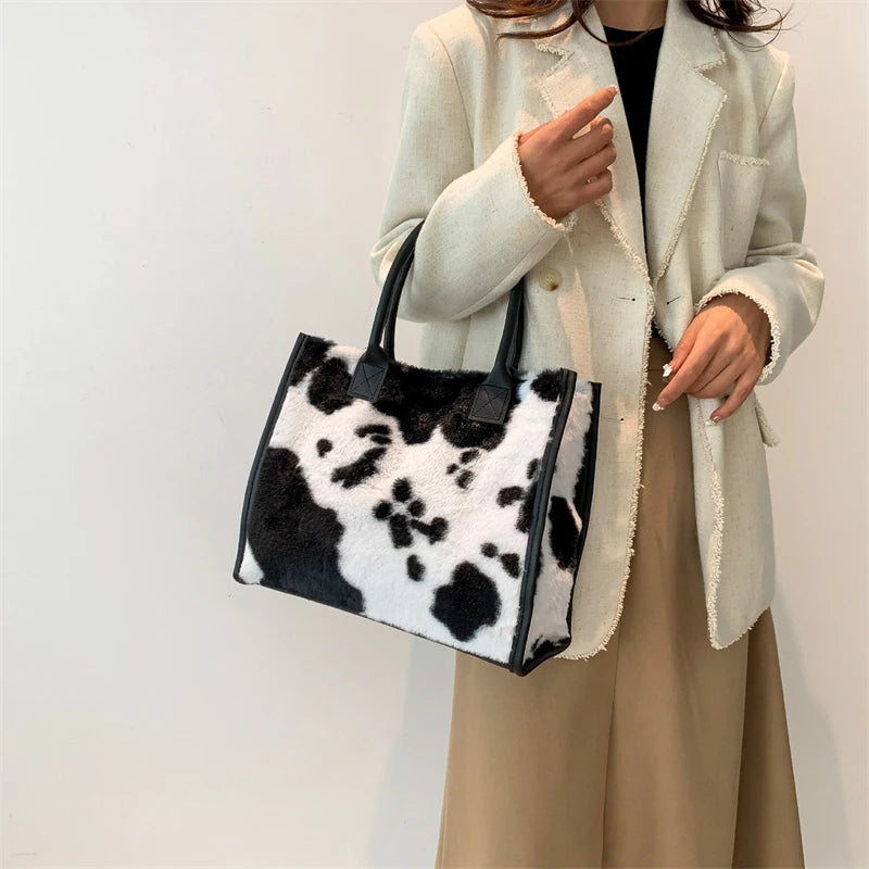 A woman holds a black and white leopard print faux fur designer shoulder tote bag in front of a light beige blazer.