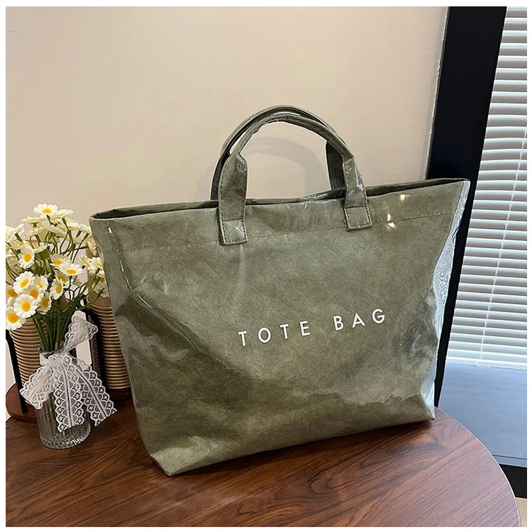 A green transparent waterproof PVC tote bag is displayed on a wooden surface next to some yellow flowers.