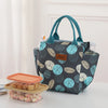 Una bolsa térmica estampada en gris oscuro y verde azulado con asas junto a un recipiente transparente de comida.