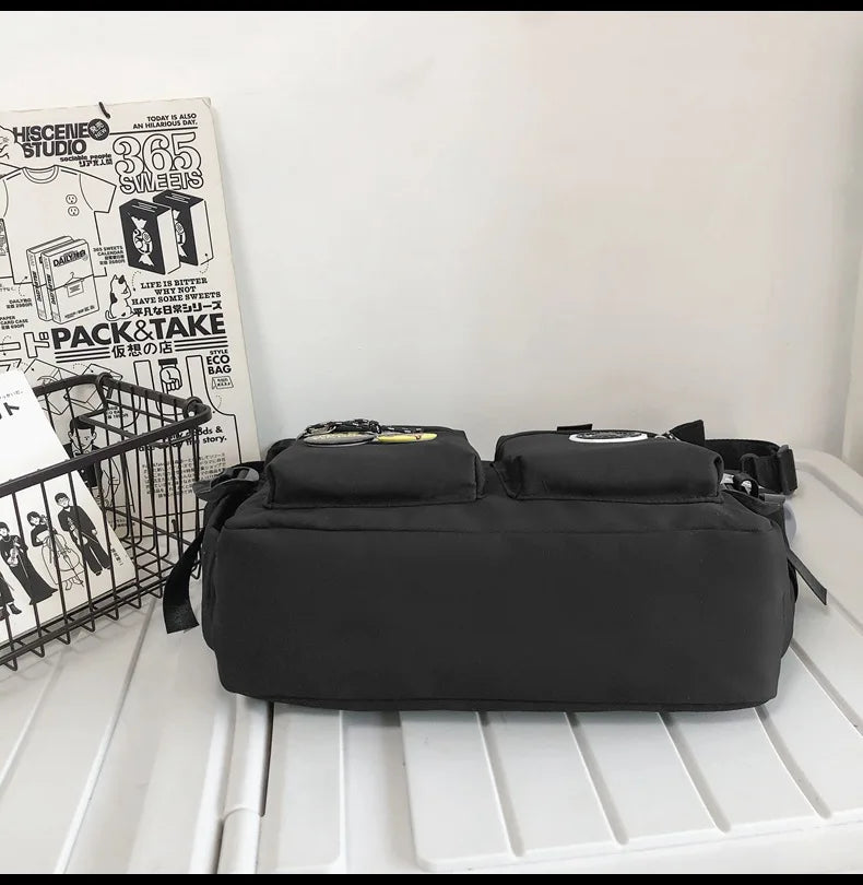 A black nylon crossbody bag is displayed on a white surface next to a stack of magazines and a wire basket.
