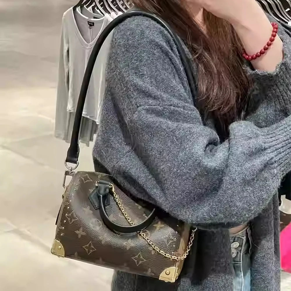 A person is holding a dark brown cowhide leather shoulder bag with a gold chain strap, worn over a gray sweater.