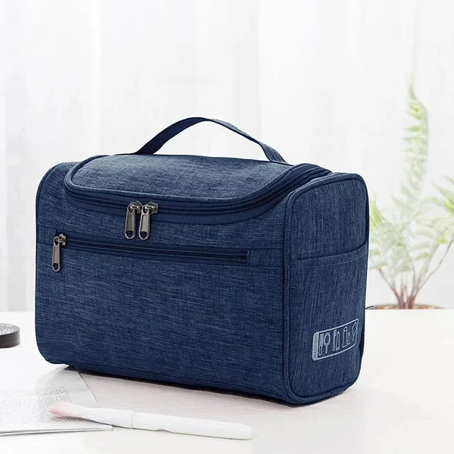A dark blue portable toiletry bag with a black handle and zippered compartments is shown on a white surface.