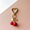 Gold heart-shaped charm with a lobster clasp and two red cherry beads on a pink leather bag.