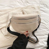 A beige soft leather crossbody bag with double zippers and a black adjustable strap resting on white bedding.