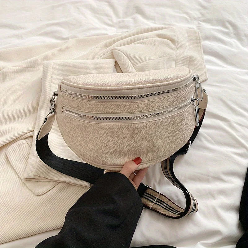 A beige soft leather crossbody bag with double zippers and a black adjustable strap resting on white bedding.