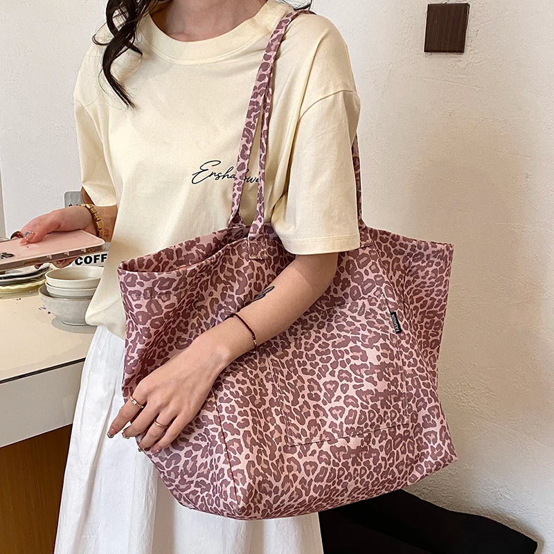 A woman holds a pink leopard print canvas tote bag against a white shirt in a bright indoor setting.