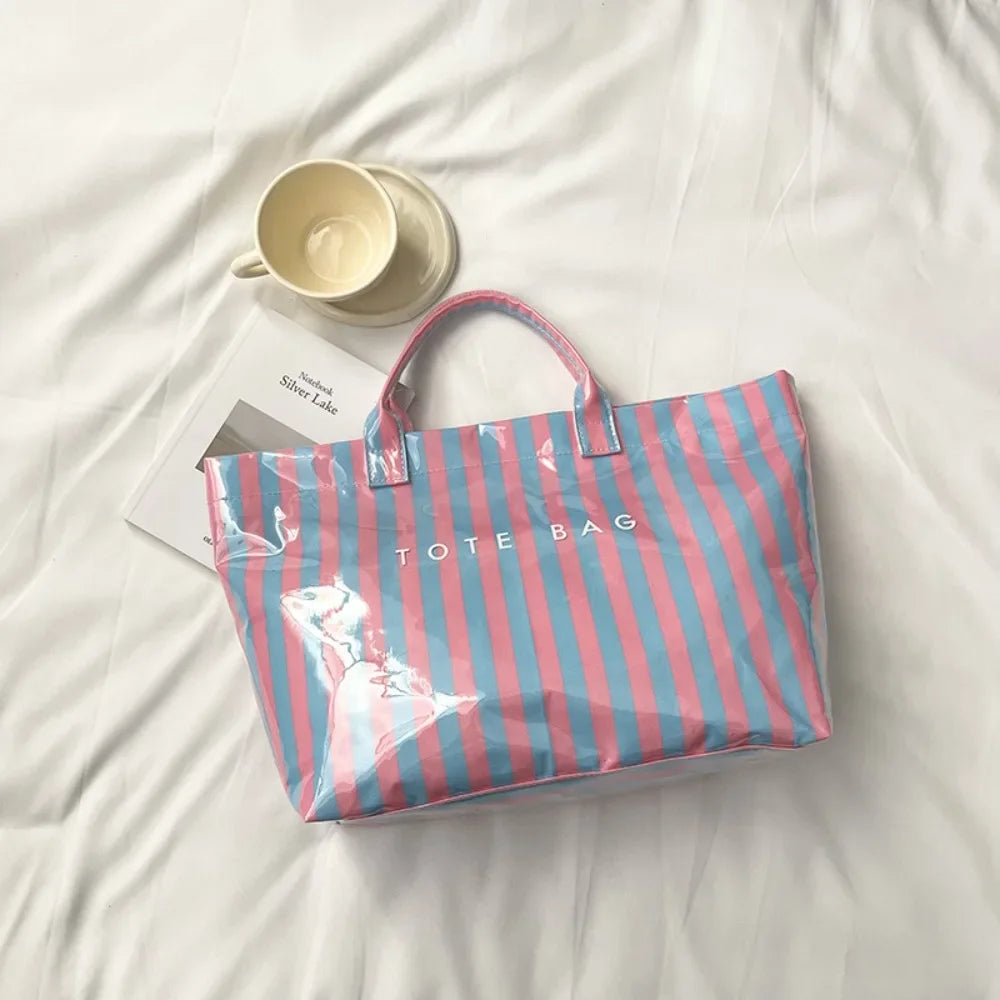 A striped transparent PVC tote bag in pink and blue stripes rests on a white sheet next to a cup.