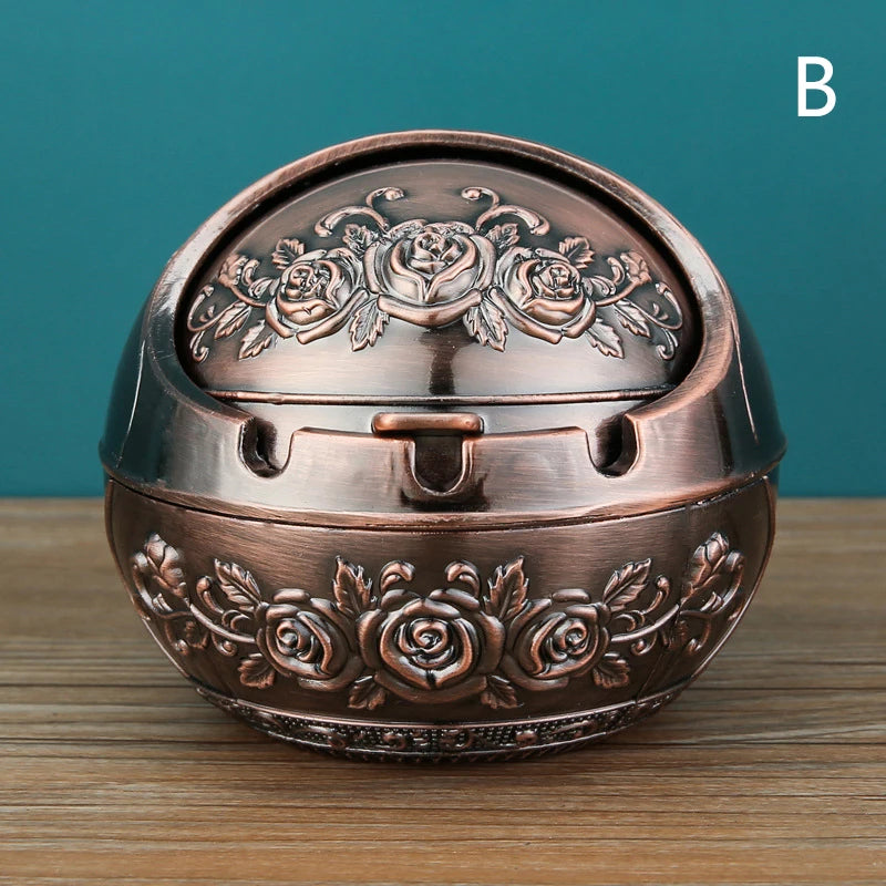 A vintage metal ashtray with a rose embossing design and a windproof lid, shown on a wooden surface.