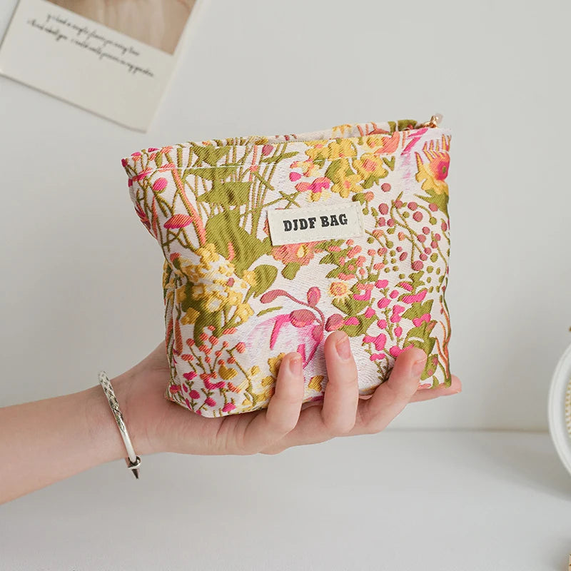 A hand holds a floral canvas makeup bag with pink and green botanical print against a white background.