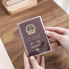 A hand holds a dark purple passport card sleeve with gold embossing on a wooden table.