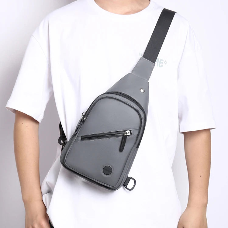 A gray adjustable crossbody sling bag worn by a person, featuring a black strap and a zippered pocket.