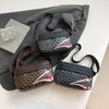 A black and gray patterned square crossbody purse resting on a dark gray fabric next to a book.