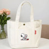 A white canvas shoulder bag featuring a Snoopy cartoon design, displayed next to pink flowers.