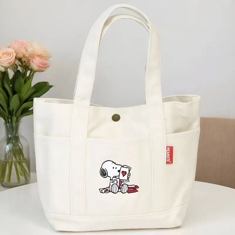 A white canvas shoulder bag featuring a Snoopy cartoon design, displayed next to pink flowers.