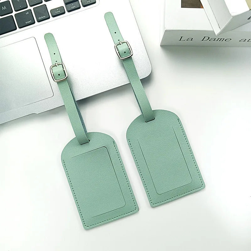 Two mint green PU leather luggage tags with silver hardware resting on a white desk next to a laptop.