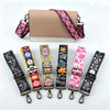 A beige leather handbag with a purple and white patterned strap is shown alongside several other colorful patterned straps.