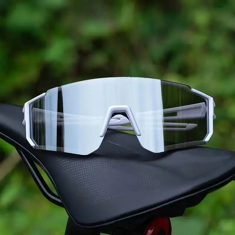 A pair of clear, lightweight oversized sunglasses resting on a black surface with blurred green foliage in the background.