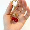 Gold heart-shaped bag charm with a lobster clasp and a dangling cherry.