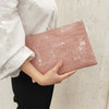 A woman holds a pink crocodile print clutch bag against a light beige wall.