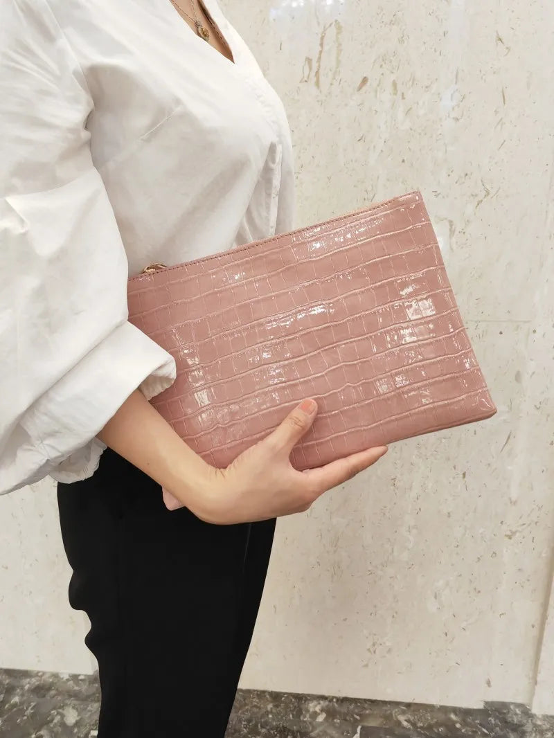 A woman holds a pink crocodile print clutch bag against a light beige wall.