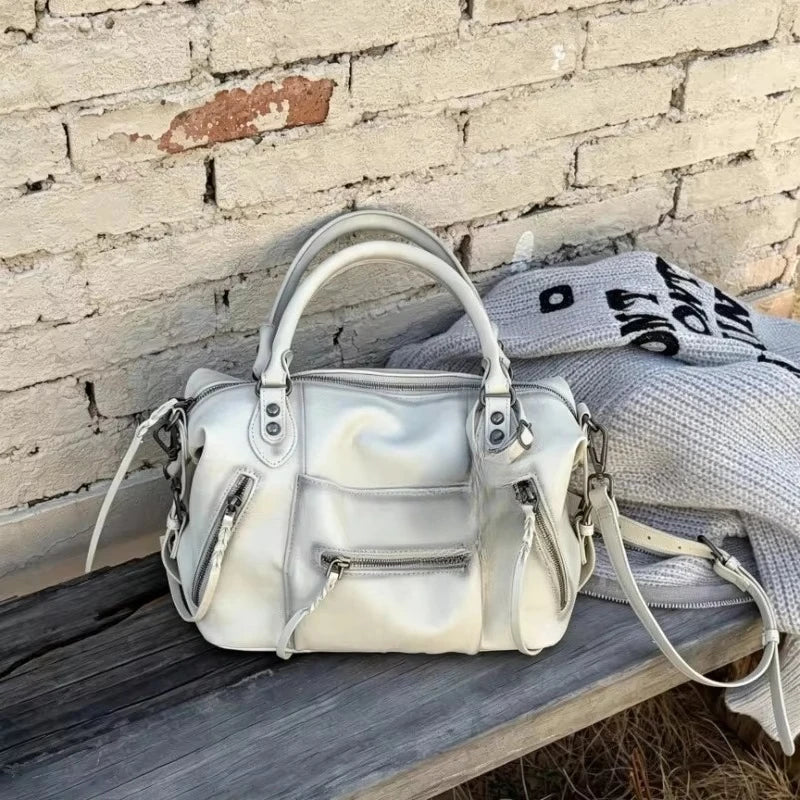 A cream-colored retro motorcycle PU leather crossbody shoulder bag resting on a wooden surface against a brick wall.